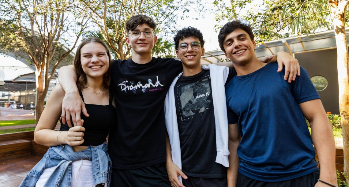 Three teenage boys and one teenage girl smile at the camera, standing side-by-side outdoors.